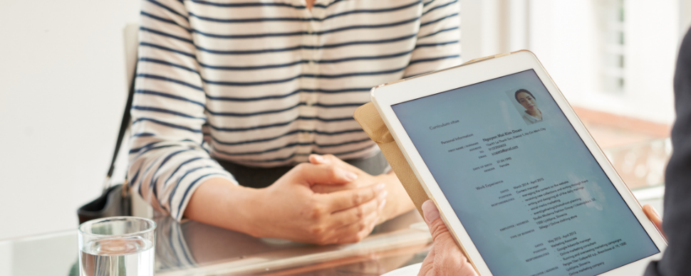 Person holding a tablet showing a resume during an interview at a glass table.