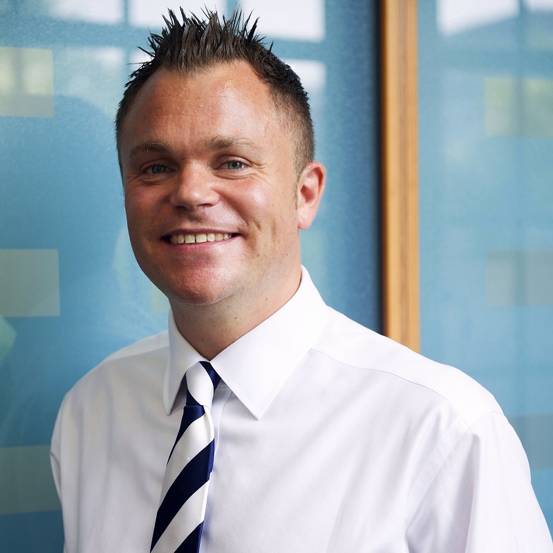 Smiling person in a white shirt and tie against a blue background.