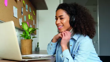 Woman with headphones smiling at laptop, working at home office desk.