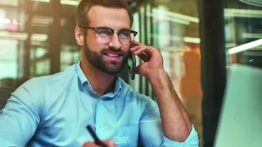 Man in a blue shirt talking on a phone and using a laptop at an office desk.