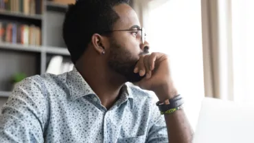 Man in patterned shirt looking thoughtfully out a window near a laptop.