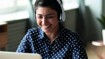 Smiling person wearing headphones and looking at a laptop in a cozy room.