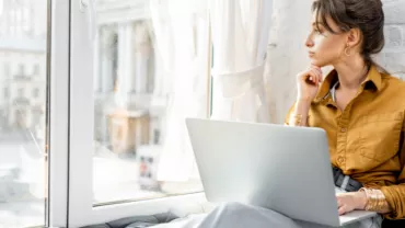 Woman sitting by a window with a laptop, looking outside thoughtfully.