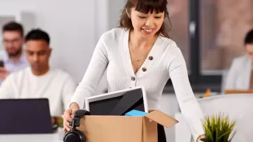 Woman unpacking office items from a box, smiling. Two colleagues work in the background.