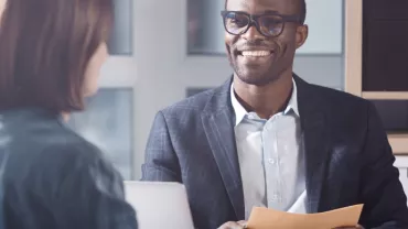 Man in glasses smiling while holding papers in an office meeting with a woman.