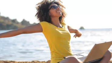 Woman in yellow shirt stretching on beach with laptop.