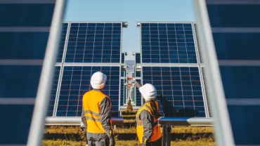 Two workers in hard hats and vests inspect large solar panels in a field.