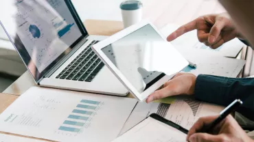 Two people reviewing financial graphs on a tablet and laptop at a desk.