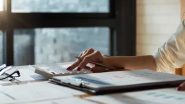 Person using a calculator at a desk with reports and glasses.