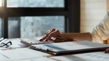 Person using a calculator at a desk with reports and glasses.