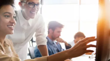 Two people smiling while working on a computer together in a bright office.