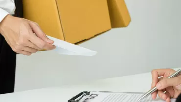 Person handing an envelope to someone signing a contract at a desk.