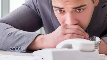 Man in a suit looks anxiously at a telephone on a desk.