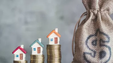 Three small house models on stacked coins beside a burlap sack with a dollar sign.