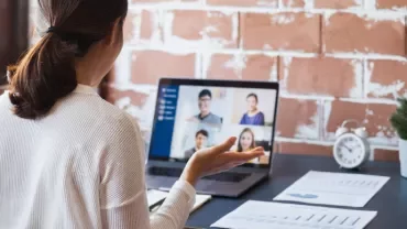 Person in a white sweater attending a video conference on a laptop at a desk with papers and a small plant.