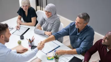A diverse group of people having a meeting around a table, with two men shaking hands.