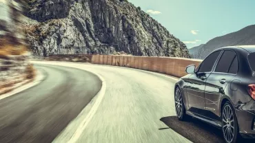A sleek black car driving on a winding mountain road under a clear sky.