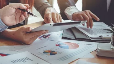 Hands holding a tablet over a table with charts and graphs.