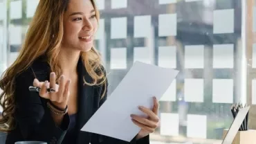 Smiling woman in an office holding a document, seated by a window with sticky notes.