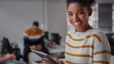 Woman in striped sweater smiling, holding a tablet in office.