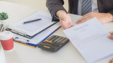 A person submits a resignation letter during a meeting with a colleague at a desk with a coffee and calculator.