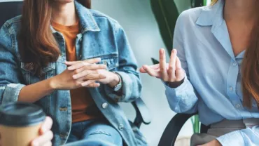 Three people in casual conversation, two women gesturing, one holding a coffee cup.