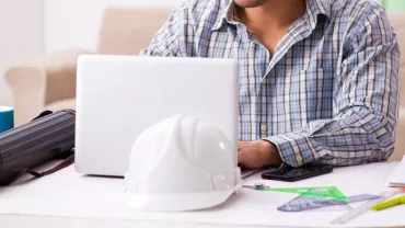 Man in plaid shirt working on a laptop with a white helmet on the table.