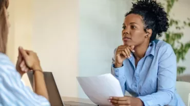 Two women having a focused discussion at a desk with a laptop and paperwork.