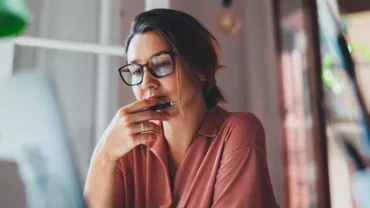 Woman in glasses working on a laptop, holding a pen to her chin and looking thoughtful.