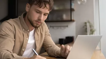 Man writing notes in front of a laptop at a home desk.