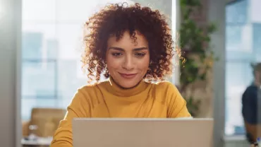 Woman with curly hair working on a laptop in a bright, plant-filled office.