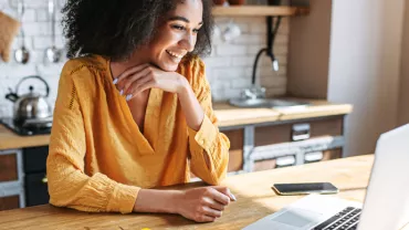 Smiling woman in yellow shirt using a laptop at a wooden table in a cozy kitchen.