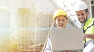 Two construction workers, a man and woman, reviewing plans on a laptop at a building site.