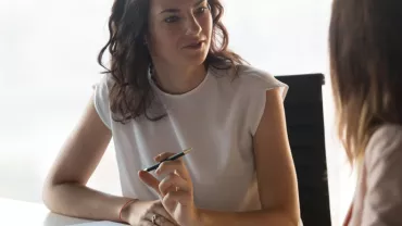 Two women having a conversation at a desk with a laptop nearby.