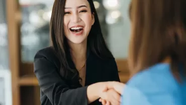 Woman in a black blazer smiling and shaking hands with another person.