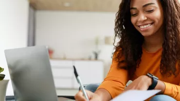 Woman in orange shirt writing on paper with laptop at table.