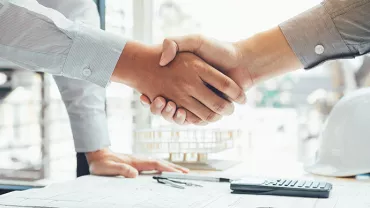 Two people shaking hands over construction blueprints.