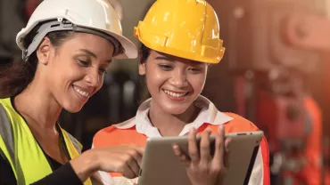 Two women in construction gear smiling while looking at a tablet.