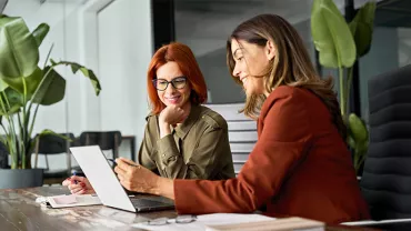 Two women discussing work at a desk in a modern office with large plants.