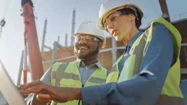 Two construction workers in safety gear review plans on a laptop at a building site.