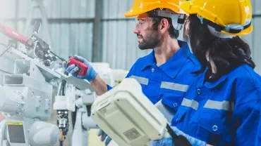 Two engineers in blue uniforms and helmets work with industrial machinery.
