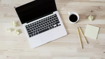 Open laptop on wooden desk with coffee, pencils, sticky notes, and crumpled paper.