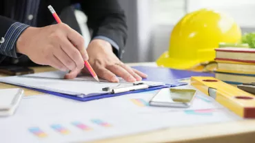 Person writing at a desk with construction tools and a yellow hard hat.