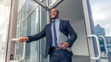 Businessman in a suit exiting a glass building, holding a laptop and smiling.