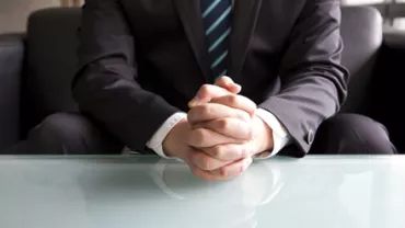 Person in suit sitting at a glass table with folded hands.
