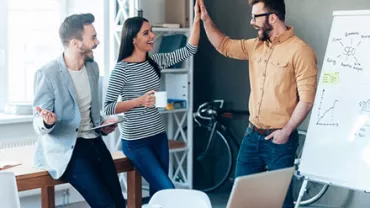 Three colleagues celebrate a success with a high-five next to a whiteboard in an office.
