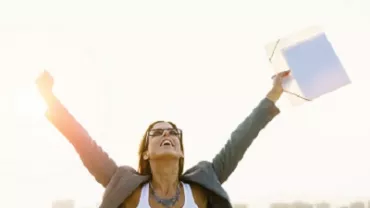 Person stands joyfully with arms raised, holding a folder against a bright sky.