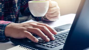 Person using a laptop while holding a coffee cup, sitting at a desk.