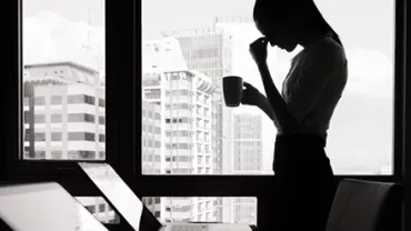 Silhouette of a person holding a cup in front of a window, with a cityscape in the background.