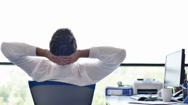 Person relaxed in office chair, hands behind head, near window.